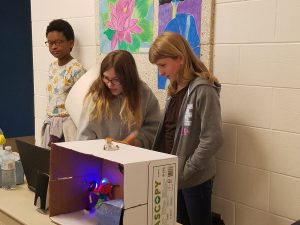Three young girls standing behind their robotic presentation. 
